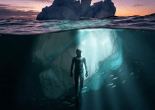 Harbor seal swimming beneath a large iceberg with a sunset visible above the water.